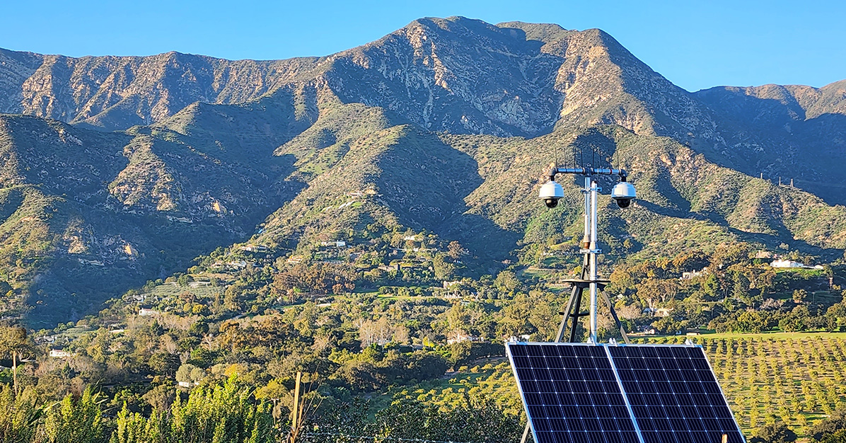 Two ALERTCalifornia cameras mounted on a large tripod with solar panels overlook Ortega Ridge near Santa Barbara, CA. The mountainous ridge is surrounded by homes and vineyards.